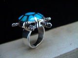 A sterling silver ring featuring a stylized turtle design with a vibrant turquoise inlay shell on a black and beige background.