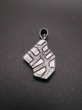 A silver pendant with a geometric, textured design featuring a central cross motif, displayed on a dark textured background.